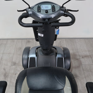 A front-facing view of a motorized three-wheel balance scooter with a digital display screen, black handlebars, and a black and gray seat, set against a light gray wall and wood-look floor.
