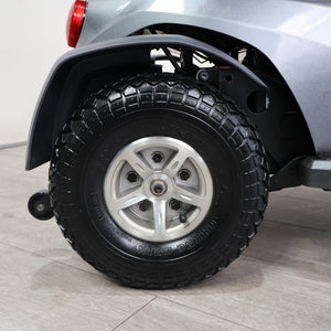 Close-up of a black all-terrain tire mounted on a silver five-spoke alloy wheel, fitted on a vehicle with a black fender, photographed indoors on a light gray tiled floor.
