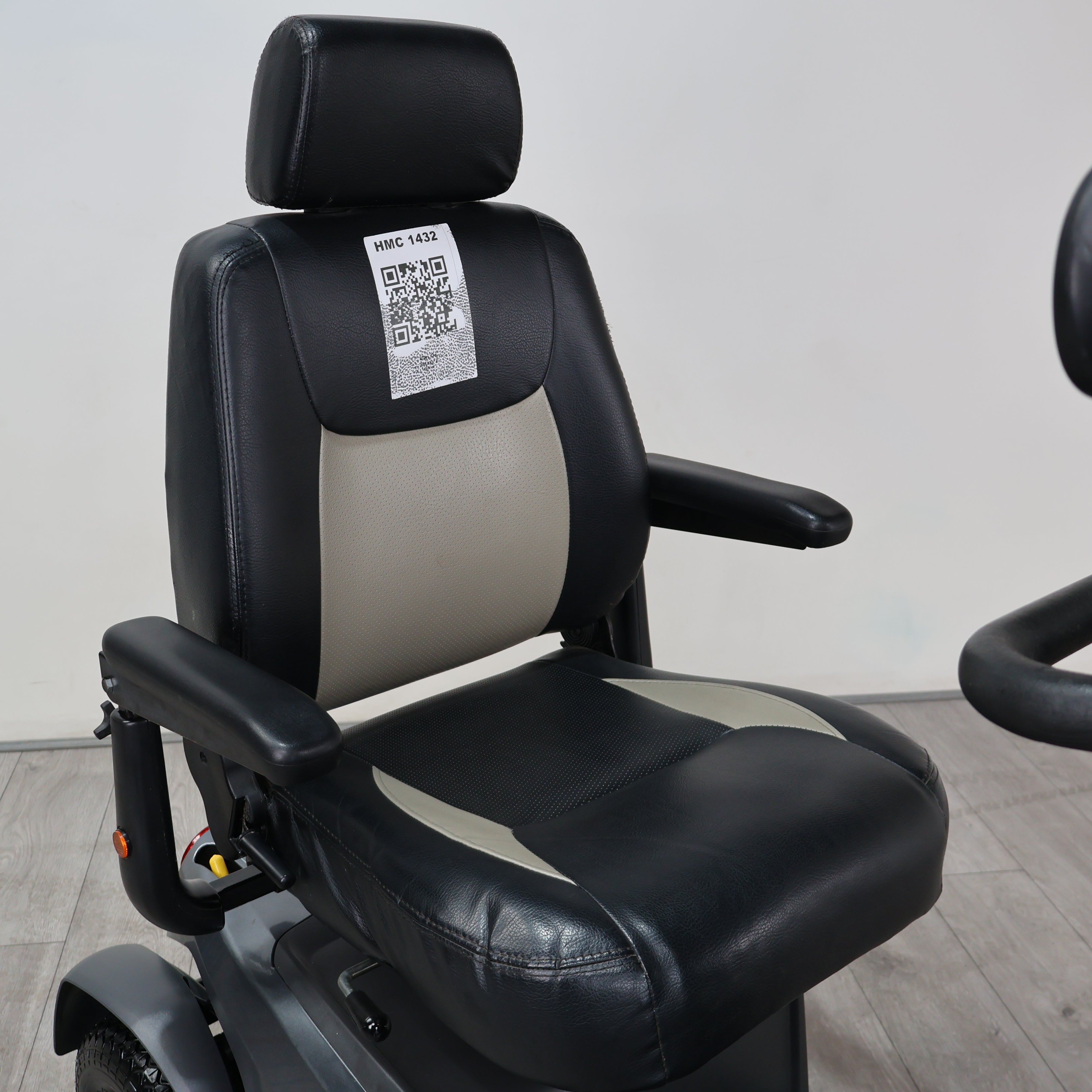 Black and beige leatherette power wheelchair seat with adjustable armrests and a headrest, shown indoors against a plain light wall.